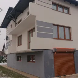 Dwupiętrowy dom z balkonami, pomalowany na biało z szarymi akcentami, brązowe okna i brama garażowa, widok z dołu na żwirowe podłoże.