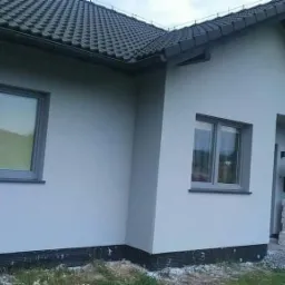 Malowanie elewacji domu jednorodzinnego na szaro; pracownik stoi na prowizorycznym rusztowaniu z białego styropianu, widoczne okna z szarymi ramami, ciemna dachówka.