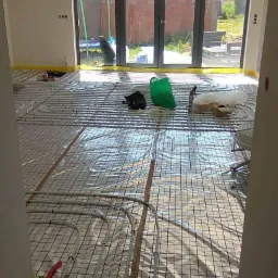 Rozłożona instalacja ogrzewania podłogowego na folii z siatką, widoczne rury mocowane do siatki, żółta taśma zabezpieczająca przy ścianach, na podłodze leżą narzędzia i torba.