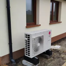 Biała zewnętrzna jednostka pompy ciepła LG Therma V, zamontowana na betonowych bloczkach przy ścianie domu z dwoma oknami, widoczny czarny system rynnowy i błotniste podłoże.