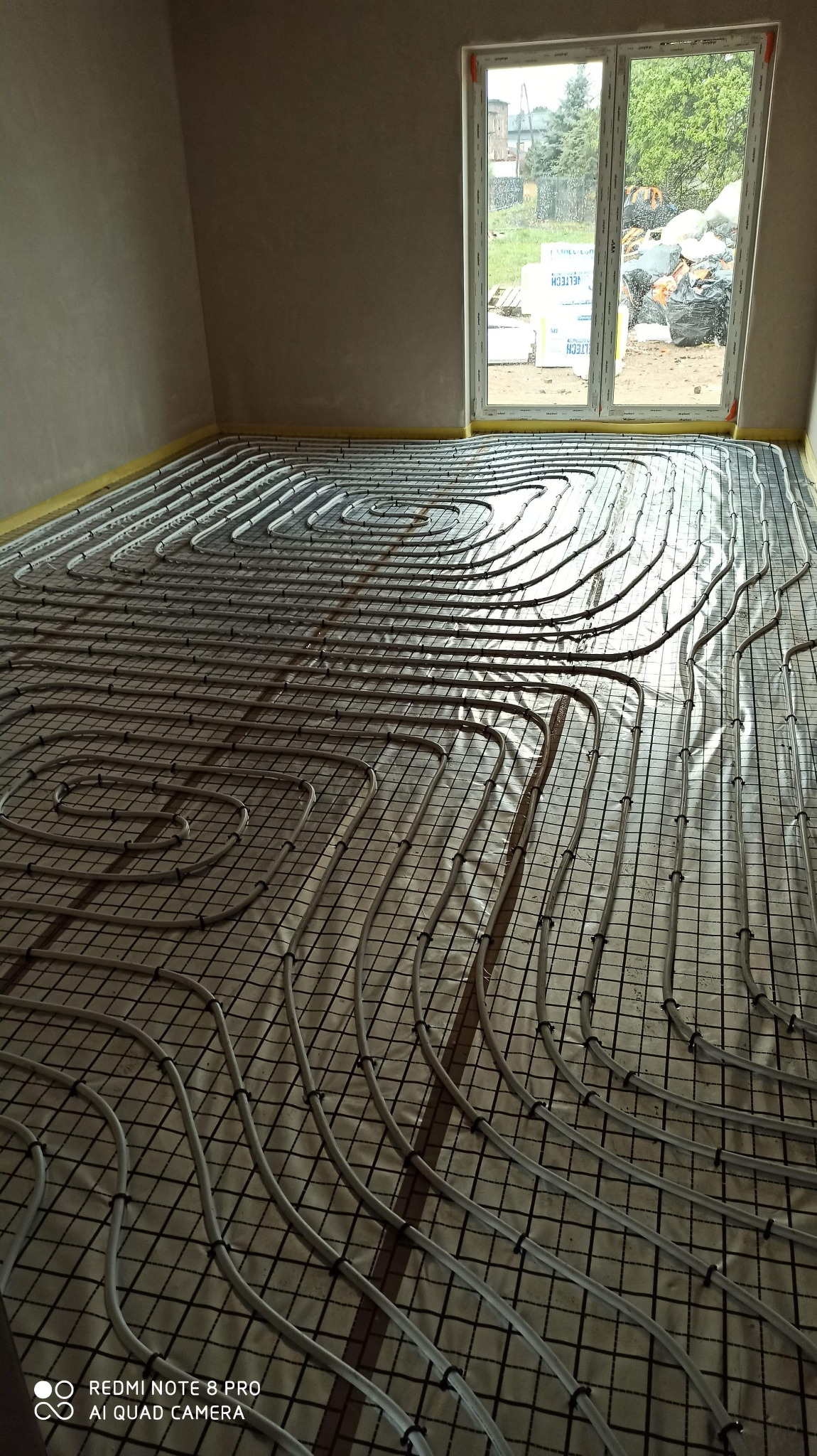 Rozłożona instalacja ogrzewania podłogowego na siatce w surowym wnętrzu, widoczne rury grzewcze i taśma izolacyjna przy ścianie, okno z widokiem na podwórko z materiałami budowlanymi.