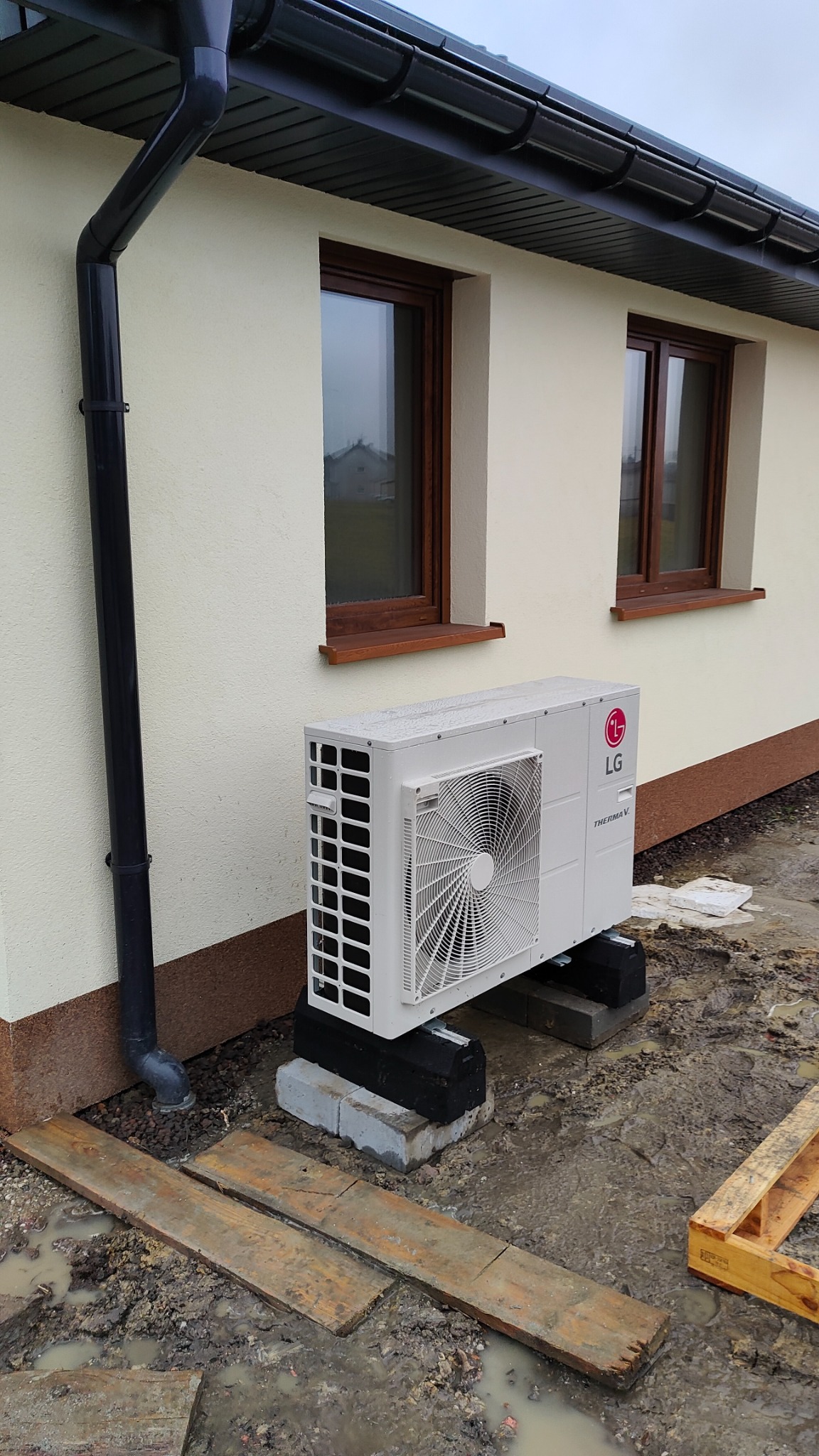 Biała zewnętrzna jednostka pompy ciepła LG Therma V, zamontowana na betonowych bloczkach przy ścianie domu z dwoma oknami, widoczny czarny system rynnowy i błotniste podłoże.
