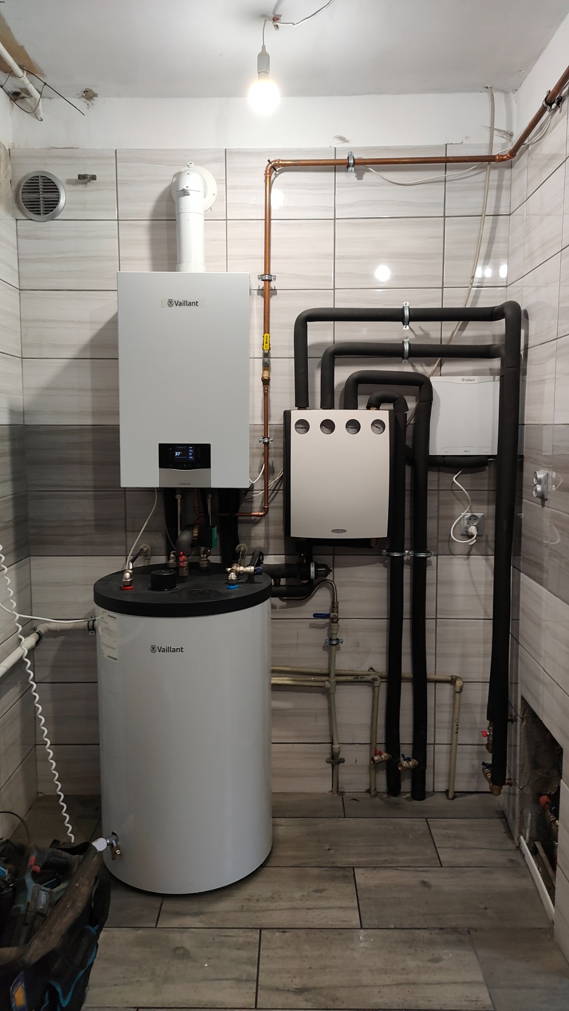 Nowoczesny, biały piec gazowy dwufunkcyjny marki Vaillant z zasobnikiem, zamontowany w pomieszczeniu wyłożonym płytkami imitującymi drewno, widoczne rury miedziane i czarne izolowane, wentylacja...
