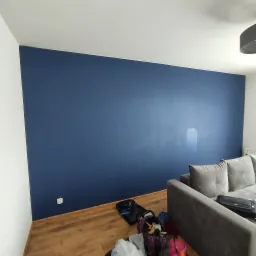 Świeżo pomalowana na granatowo ściana w salonie, widoczny fragment szarej sofy, podłoga z paneli i torby z akcesoriami malarskimi.