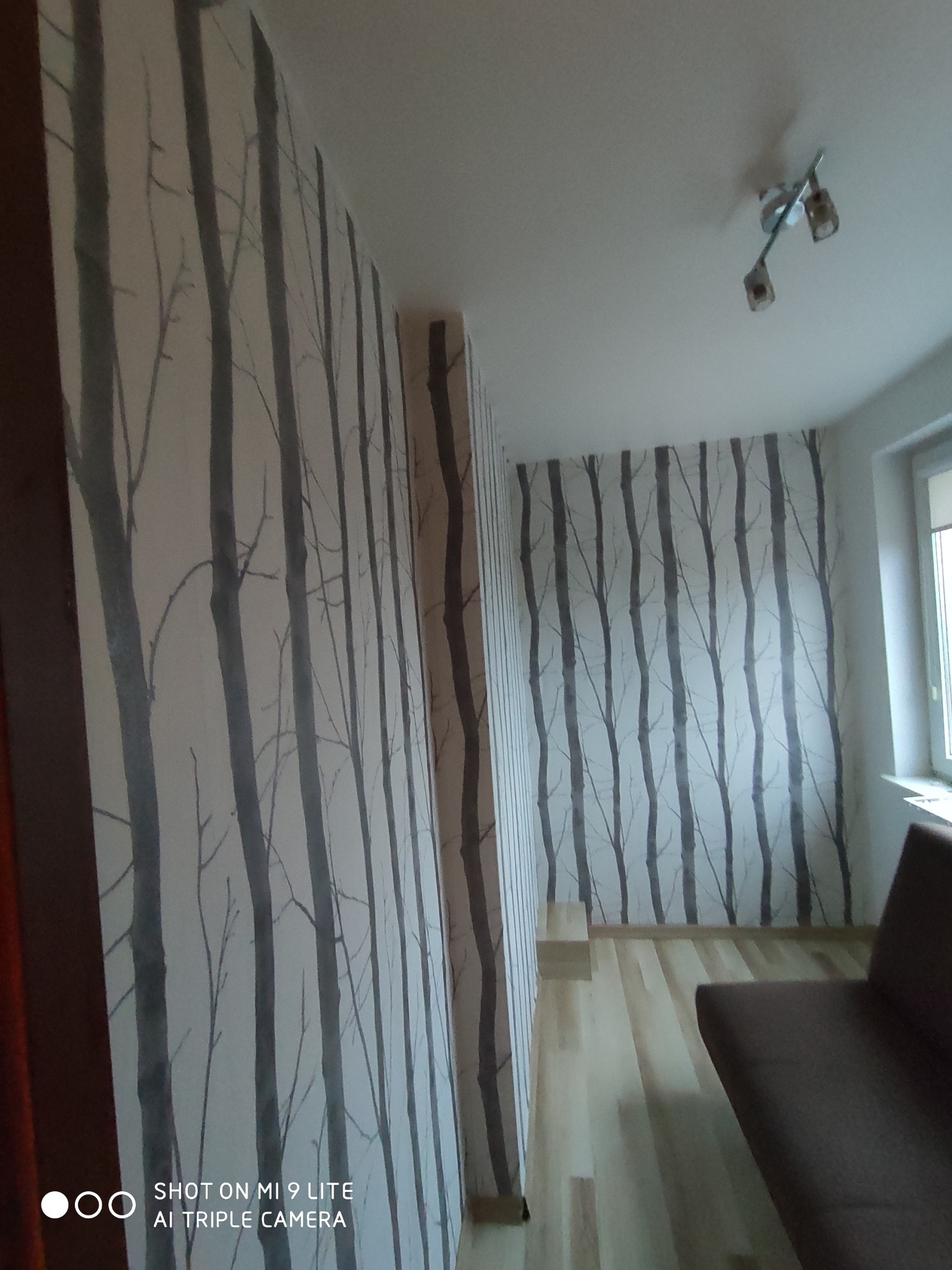 Wnętrze pokoju z tapetą w motywie drzew, jasną podłogą i częściowo widoczną sofą.