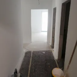 Korytarz w trakcie remontu, ściany pomalowane na biało, podłoga zabezpieczona matą ochronną z żółtym pasem, widoczny agregat malarski Makita i żółte wiadro z resztkami farby.