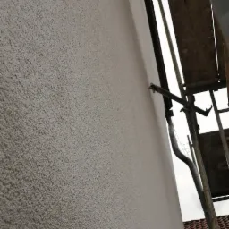 Ściana zewnętrzna budynku pokryta nowym, jasnoszarym tynkiem strukturalnym typu baranek, widoczne rusztowanie w trakcie prac.