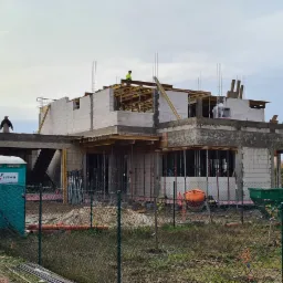 Budowa domu w stanie surowym otwartym: widoczne ściany z bloczków, elementy szalunków, stalowe pręty zbrojeniowe, pracownicy na dachu i na stropie, przenośna toaleta na placu budowy.