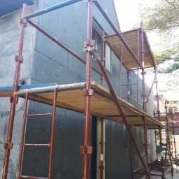 Dom w trakcie ocieplania, widoczne rusztowanie i ściana pokryta ciemnym materiałem izolacyjnym, prawdopodobnie styropianem grafitowym.