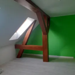 Poddasze z drewnianą konstrukcją nośną, pomalowaną na brązowo, białą skośną ścianą z oknem dachowym i intensywnie zieloną ścianą boczną, na podłodze panele imitujące drewno.