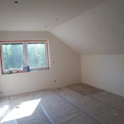 Poddasze ze skośnym sufitem po pracach przygotowawczych: wygładzone ściany, zakryta podłoga, okno z widokiem na zieleń.