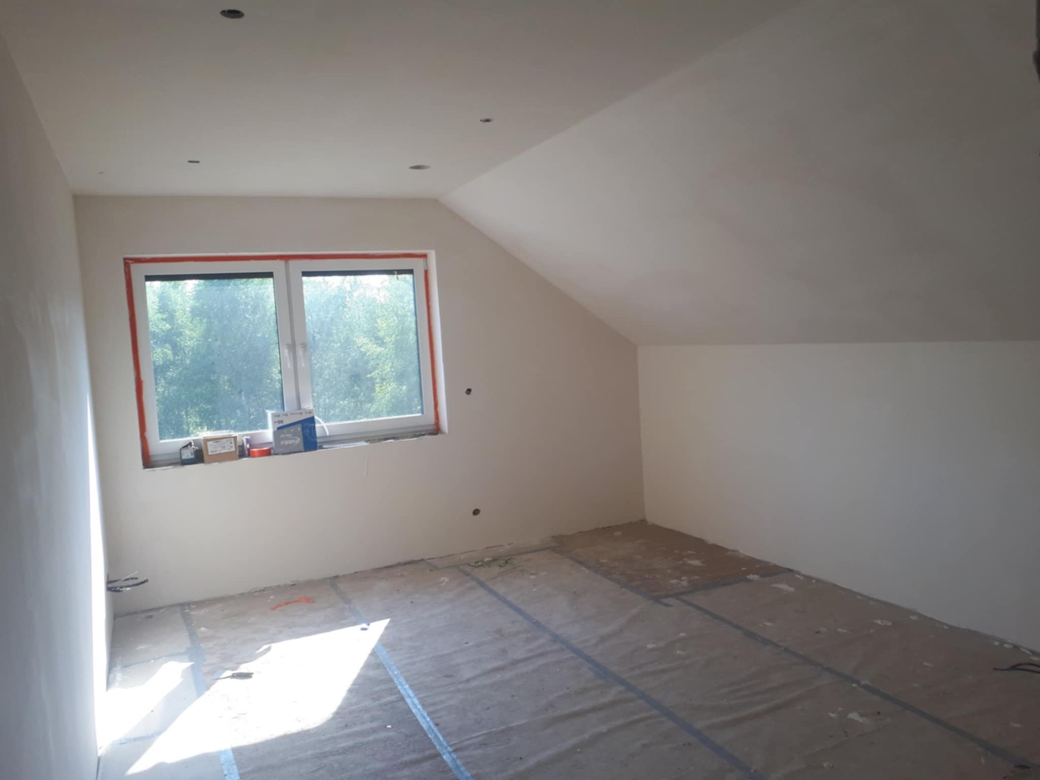 Poddasze ze skośnym sufitem po pracach przygotowawczych: wygładzone ściany, zakryta podłoga, okno z widokiem na zieleń.