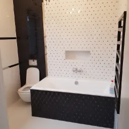 Elegancka łazienka po remoncie: biała wanna z czarnym panelem dekoracyjnym, toaleta, grzejnik drabinkowy i biało-czarne płytki z akcentami w postaci kryształków oraz ozdobny czarny żyrandol.