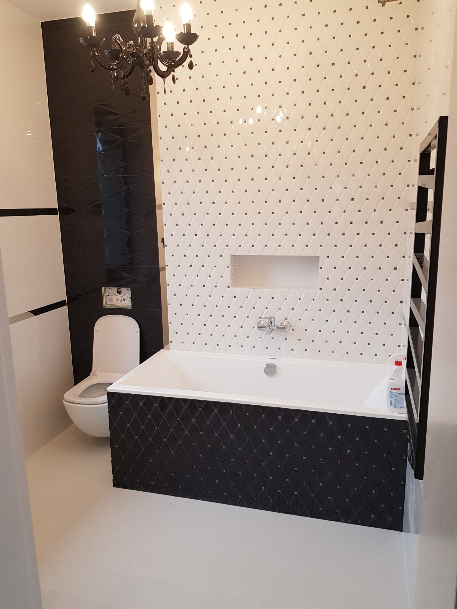 Elegancka łazienka po remoncie: biała wanna z czarnym panelem dekoracyjnym, toaleta, grzejnik drabinkowy i biało-czarne płytki z akcentami w postaci kryształków oraz ozdobny czarny żyrandol.