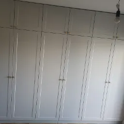 Wysoka, szara szafa z podwójnymi drzwiami i złotymi gałkami, sięgająca sufitu, z żarówką na kablu w tle.