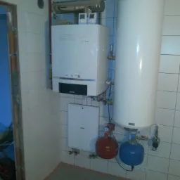 Biały piec gazowy Buderus z podłączonym zasobnikiem ciepłej wody, naczynia wzbiorcze w kolorze czerwonym i niebieskim, instalacja w pomieszczeniu wyłożonym białymi płytkami.