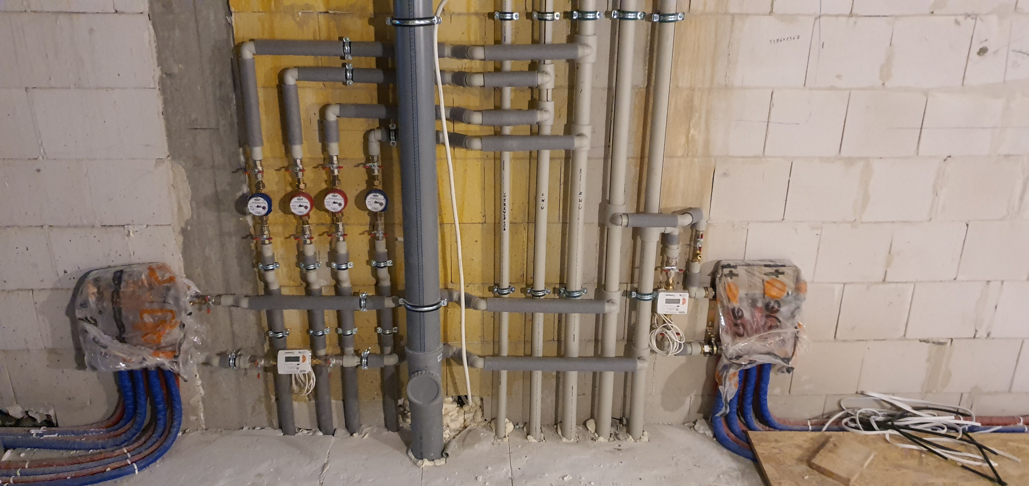Wewnętrzna instalacja wodna i grzewcza w surowym pomieszczeniu: rury z izolacją, liczniki wody, przewody doprowadzające pod folią ochronną, widoczne połączenia i mocowania.