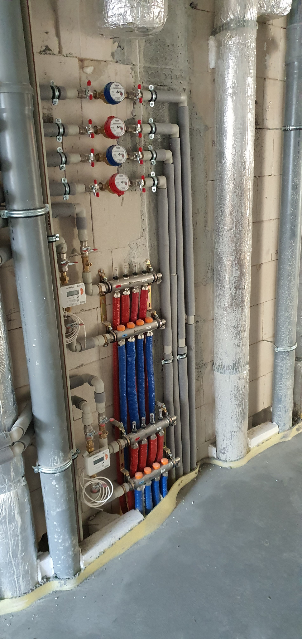 Rozdzielacz ogrzewania podłogowego z rurami w kolorze czerwonym i niebieskim, liczniki ciepła z czerwonymi i niebieskimi wskazówkami zamontowane na ścianie z cegły, rury izolowane termicznie.