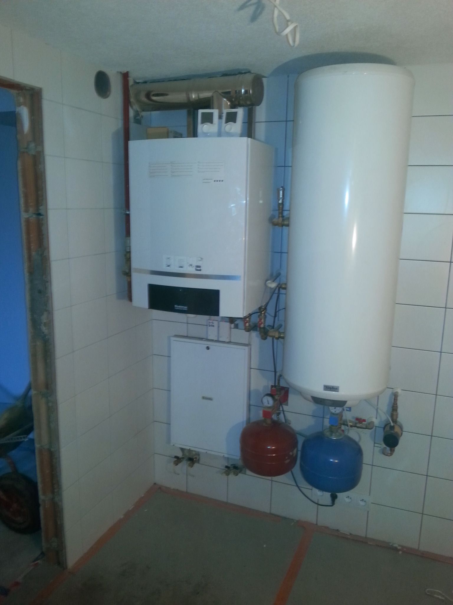 Biały piec gazowy Buderus z podłączonym zasobnikiem ciepłej wody, naczynia wzbiorcze w kolorze czerwonym i niebieskim, instalacja w pomieszczeniu wyłożonym białymi płytkami.