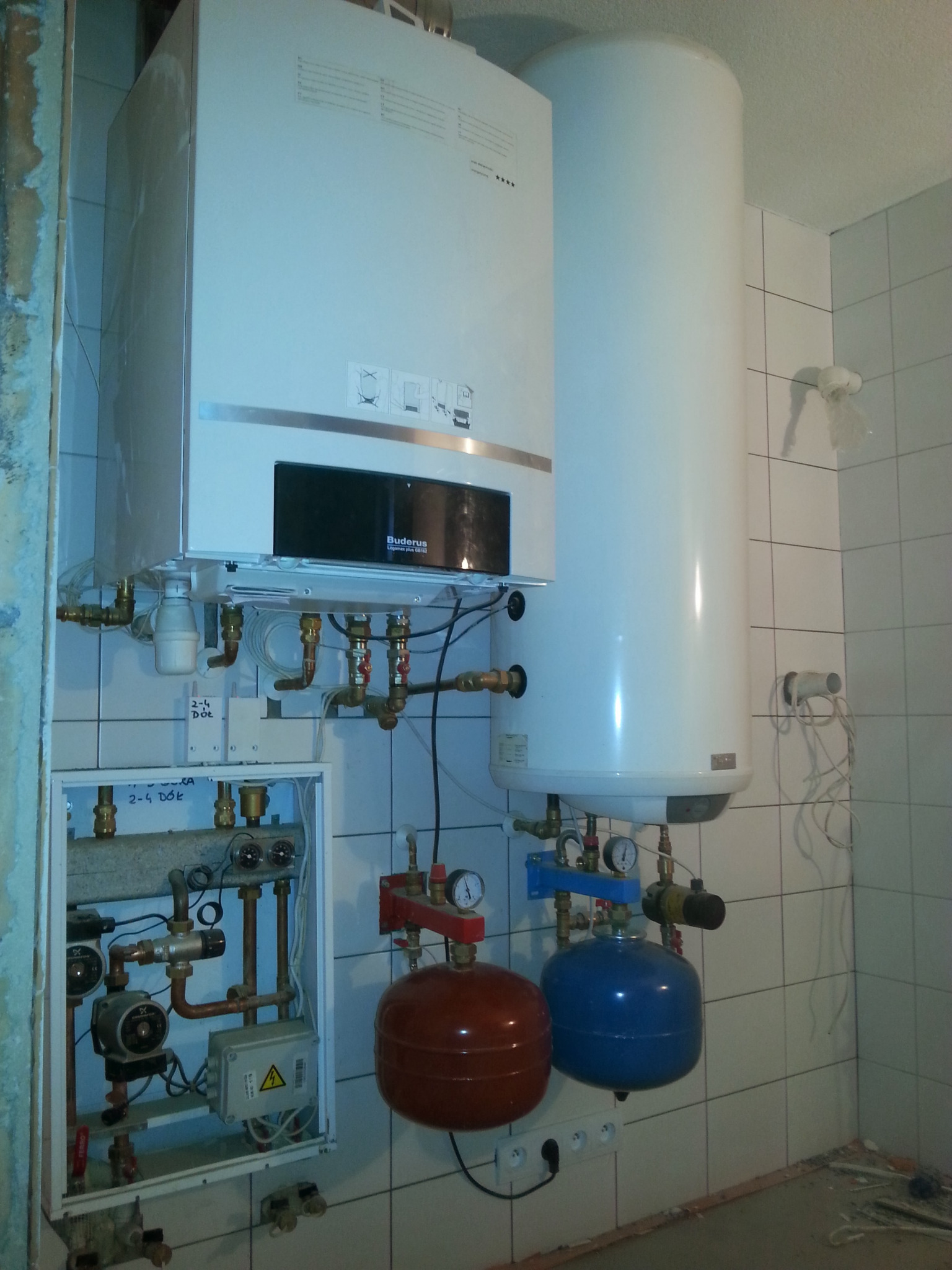 Biały piec gazowy marki Buderus Legamax plus GB192, obok biały zasobnik ciepłej wody, poniżej czerwony i niebieski zbiornik przeponowy, instalacja z rur miedzianych w trakcie podłączania...