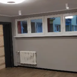 Wnętrze pokoju po odświeżeniu, ściany pomalowane na szaro, białe okna z widokiem na budynki, grzejnik żeberkowy pod oknem, widoczne lampy sufitowe