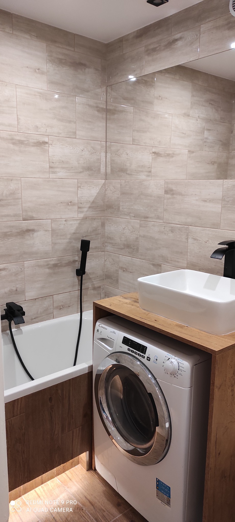 Nowoczesna łazienka z beżowymi płytkami imitującymi drewno, białą wanną z czarną armaturą, białą umywalką nablatową na drewnianym blacie oraz pralką zabudowaną pod blatem.
