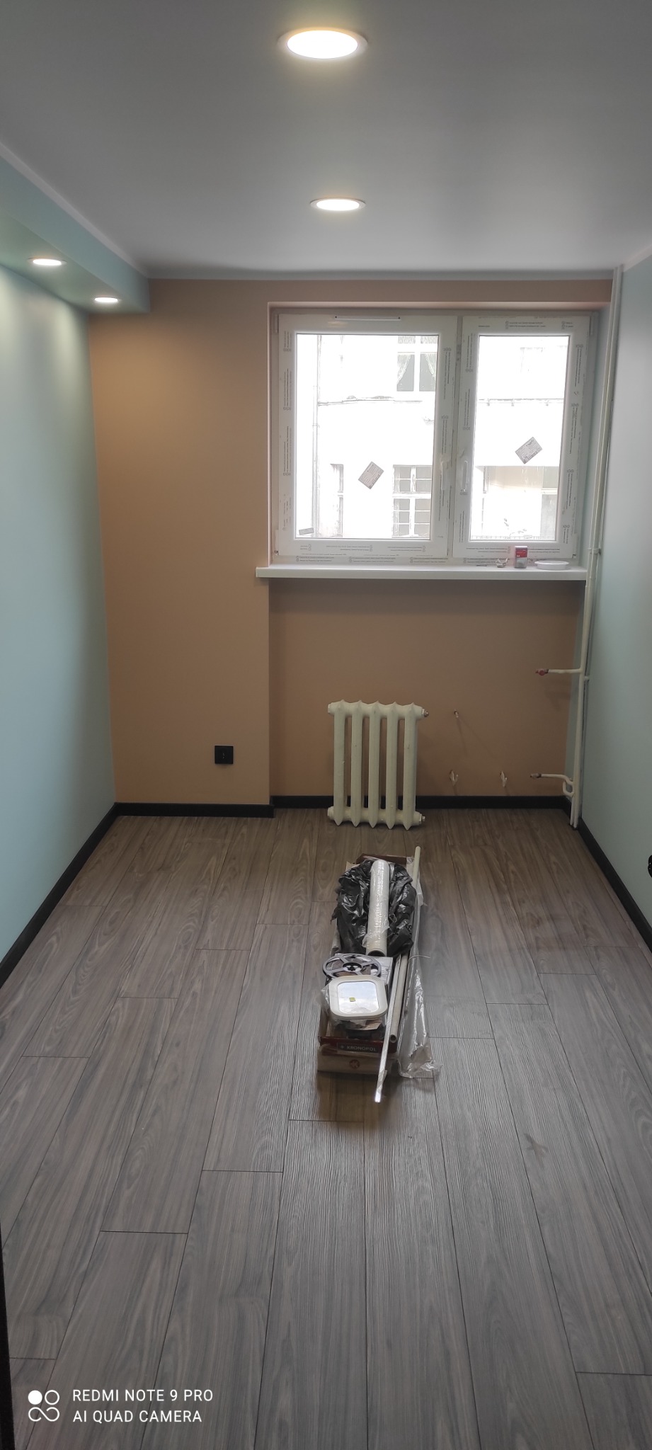 Pusty pokój po odświeżeniu z nową podłogą, pomalowanymi ścianami w dwóch odcieniach i przygotowanymi materiałami do dalszych prac w centralnej części pomieszczenia.