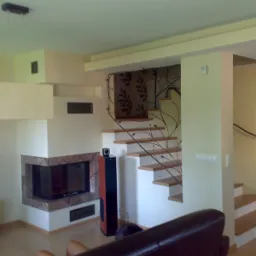 Salon z kominkiem obudowanym kamieniem i drewnianymi schodami z metalową balustradą o motywie roślinnym.