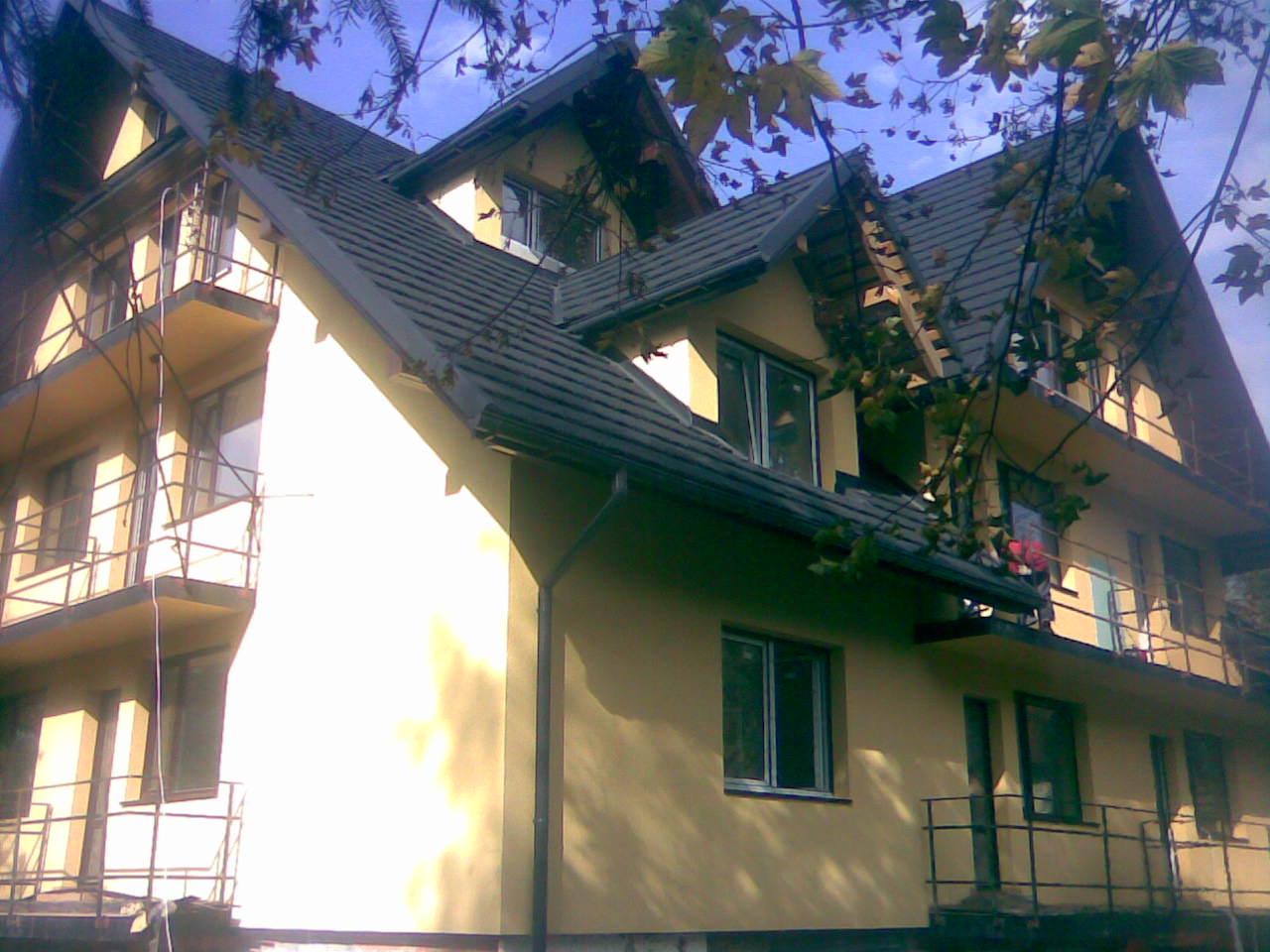 Żółty dom z ciemną dachówką widziany z dołu, częściowo zasłonięty gałęziami drzewa.