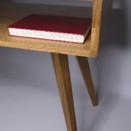 Drewniana szafka nocna z półką i skośnymi nogami w stylu retro, na półce leży czerwony notatnik.