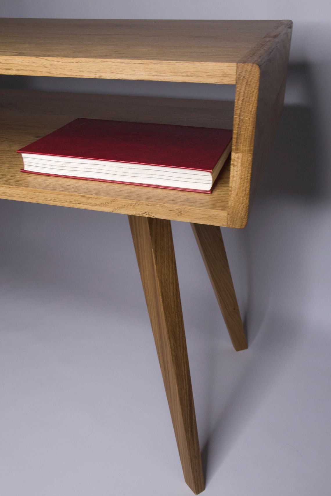 Drewniana szafka nocna z półką i skośnymi nogami w stylu retro, na półce leży czerwony notatnik.