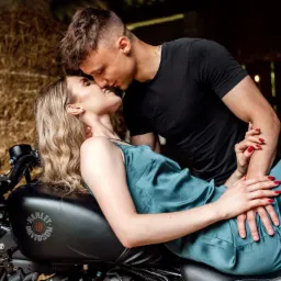 Zakochana para na motocyklu Harley-Davidson w rustykalnym otoczeniu stodoły, kobieta w jedwabnej sukience i mężczyzna w czarnym t-shircie.