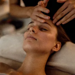 Masaż głowy i twarzy wykonywany na młodej kobiecie leżącej na jasnym posłaniu, z delikatnym oświetleniem świec w tle.