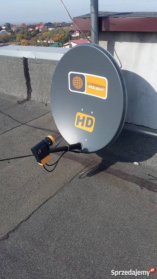 Szara antena satelitarna Cyfrowy Polsat HD zamontowana na płaskim dachu pokrytym papą, widok na miasto w tle.