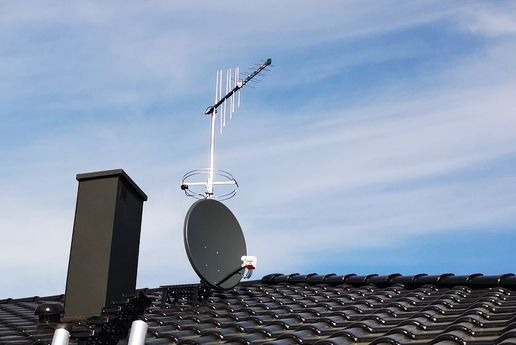 Antena telewizyjna i satelitarna zamontowane na dachu domu z czarną dachówką, widoczne na tle błękitnego nieba z delikatnymi chmurami, obok komin wentylacyjny.