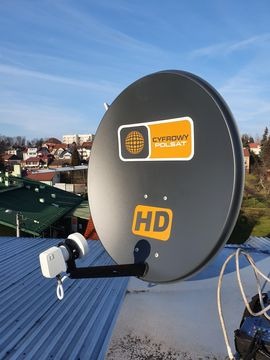 Szara antena satelitarna Cyfrowy Polsat HD zamontowana na metalowym dachu budynku z widokiem na miasto i błękitne niebo.