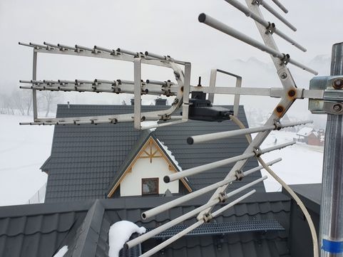Antena telewizyjna zamontowana na dachu domu z szarą dachówką w zimowej scenerii górskiej, widoczne elementy mocujące i przewody.