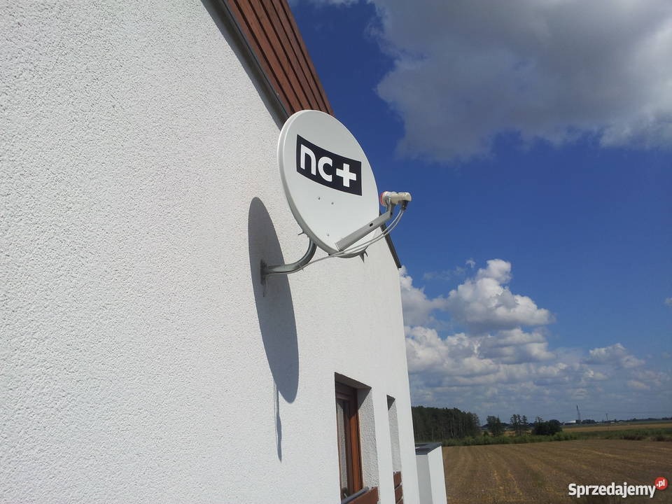 Biała antena satelitarna z logo nc+ zamontowana na ścianie budynku z teksturą baranka, na tle błękitnego nieba z chmurami i polami uprawnymi.