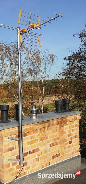 Żółto-srebrna antena telewizyjna zamontowana na kominie z cegły, widok z dołu na tle błękitnego nieba i drzew.