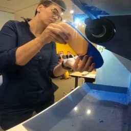 Kobieta w okularach, w niebieskiej koszuli, przygotowuje ortopedyczną wkładkę do butów w specjalistycznej maszynie. W tle siedzą osoby obserwujące proces.