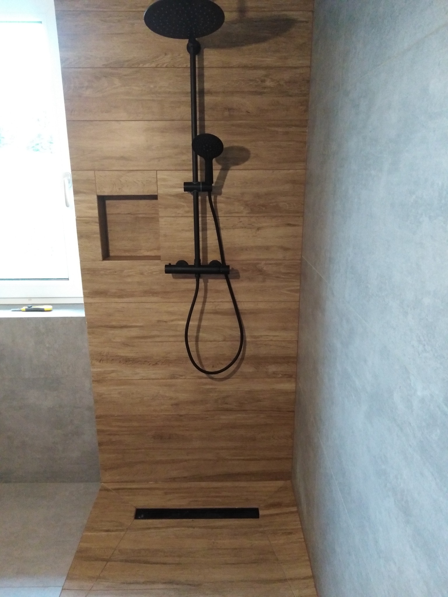 Nowoczesny prysznic z czarną armaturą i odpływem liniowym, obłożony płytkami imitującymi drewno, z wnęką w ścianie na kosmetyki.