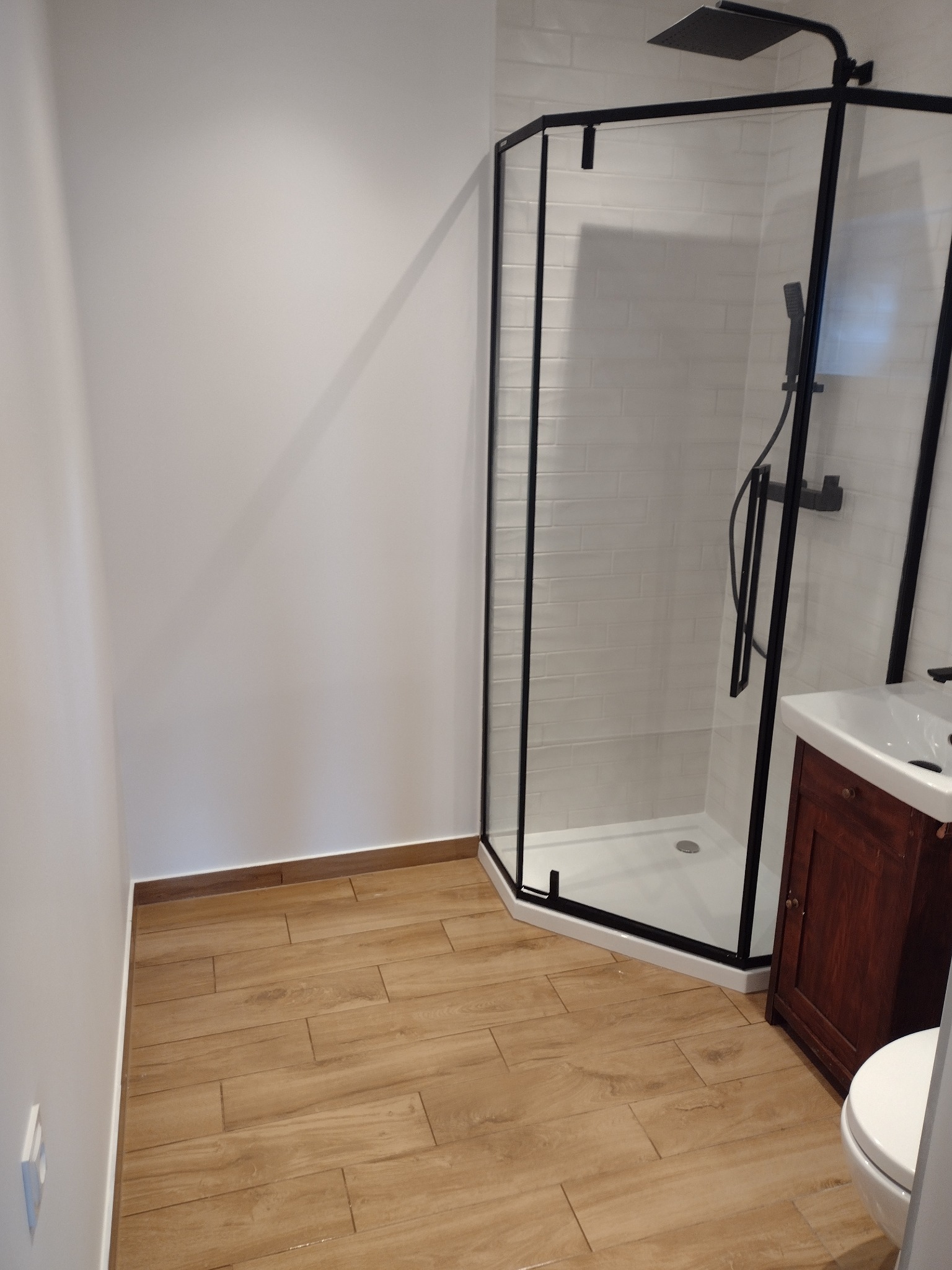 Nowoczesna łazienka z kabiną prysznicową typu walk-in z czarnymi profilami, drewnopodobną podłogą i białą armaturą.