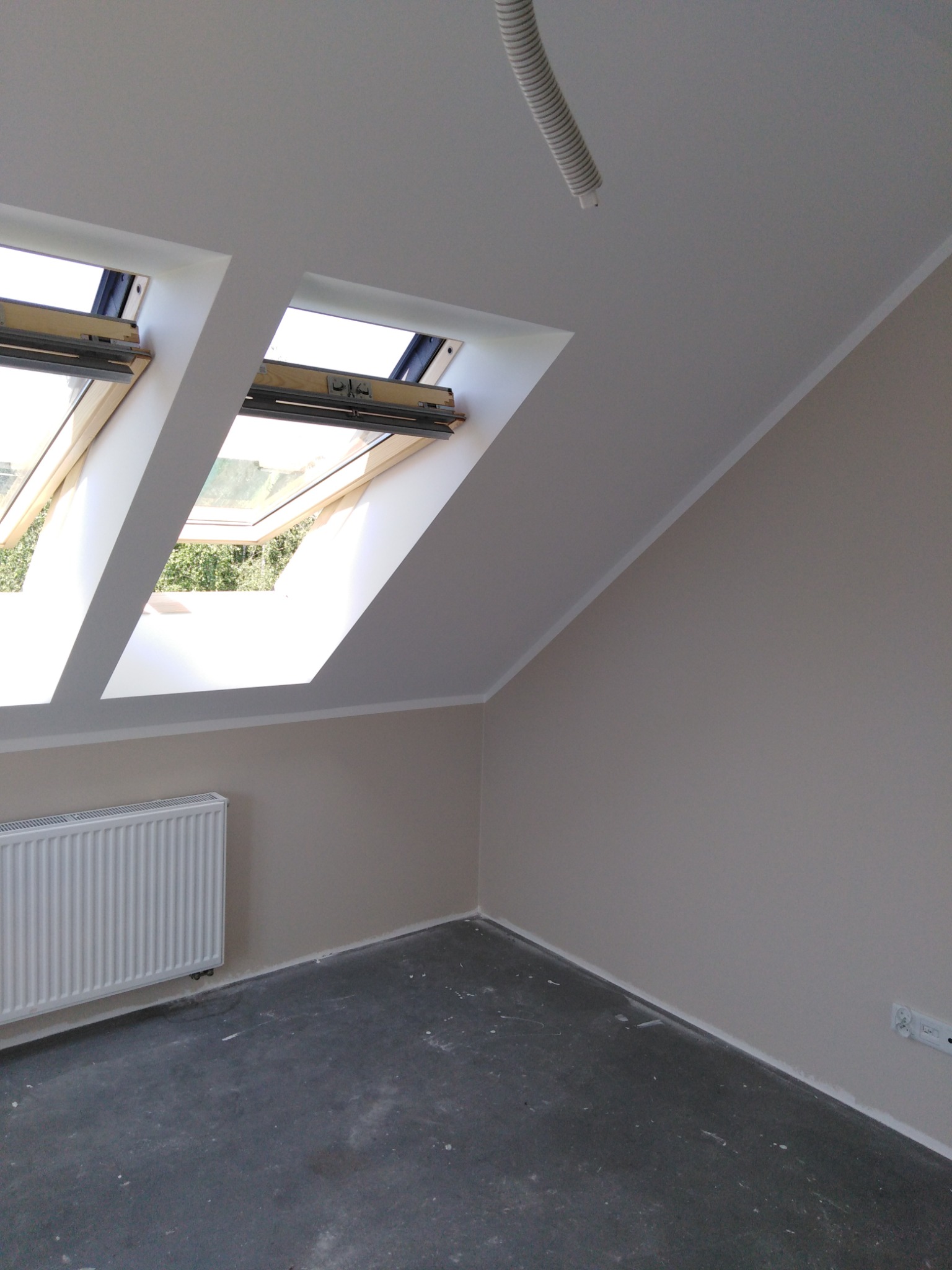 Pokój na poddaszu po remoncie, ściany pomalowane na jasny beż, dwa okna dachowe w białych obramowaniach, szary betonowy podkład podłogowy, biały grzejnik żeberkowy.