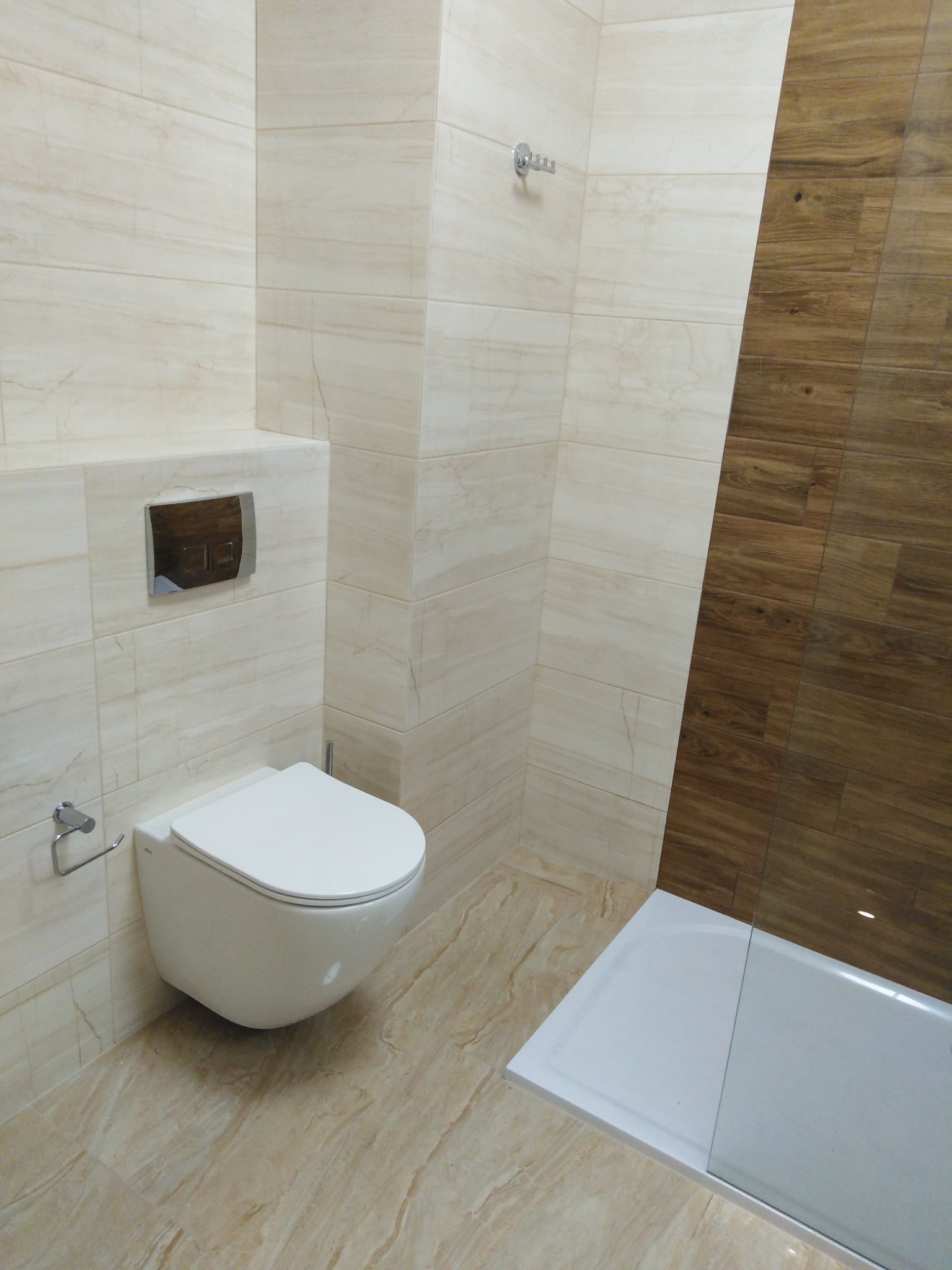 Nowoczesna łazienka z beżowymi płytkami imitującymi marmur, wisząca toaleta z chromowanym przyciskiem spłukiwania i kabina prysznicowa z drewnopodobnymi panelami na jednej ze ścian.