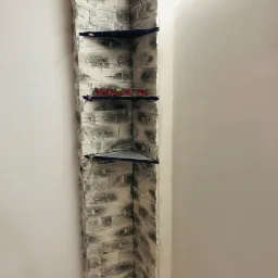 Wykończenie narożnika ściany imitacją kamienia w odcieniach szarości, z zamontowanymi półkami tymczasowo podpartymi poziomnicą, widoczne surowe ściany.