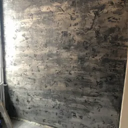 Ściana z nałożoną szarą, dekoracyjną strukturą, imitującą beton, z widocznymi śladami pędzla i nierównościami, na podłodze szare płytki, w rogu fragment drabiny.