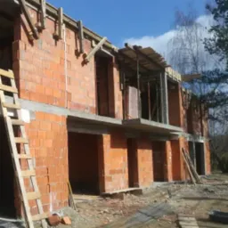 Wznoszenie dwukondygnacyjnego budynku z czerwonej cegły, widoczne drewniane szalunki i drabina oparta o ścianę, surowy stan.