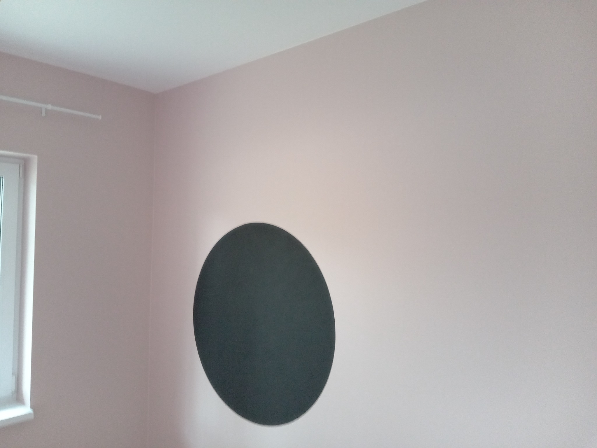 Ściana pomalowana na delikatny róż z ciemnoszarym, okrągłym akcentem dekoracyjnym, widoczne okno i fragment sufitu.