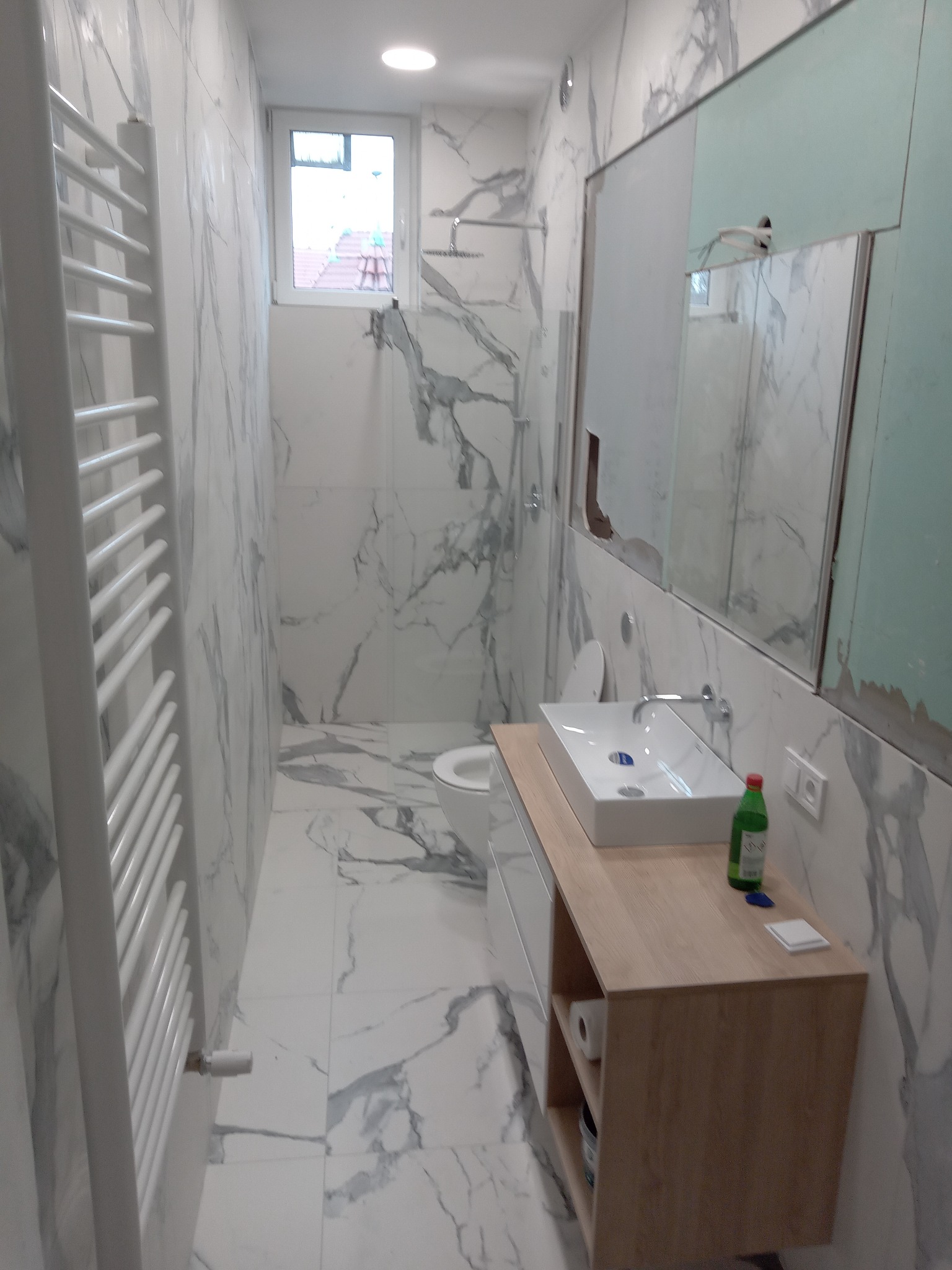 Wnętrze łazienki z marmurowymi płytkami, grzejnikiem drabinkowym, umywalką nablatową na drewnianej szafce, lustrem w trakcie montażu i widoczną instalacją hydrauliczną.