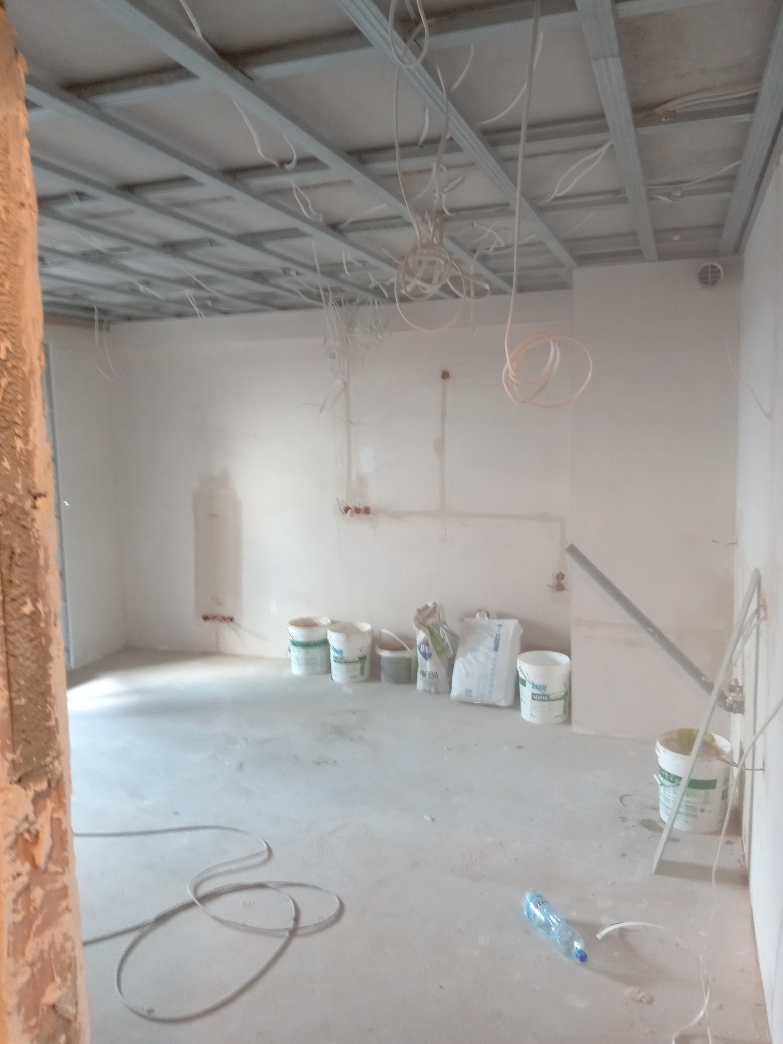 Wnętrze w trakcie prac wykończeniowych: widoczna konstrukcja sufitu podwieszanego, ściany po wstępnym nałożeniu gładzi, worki z zaprawą i wiadra z farbą przy ścianie, oraz butelka wody na podłodze.